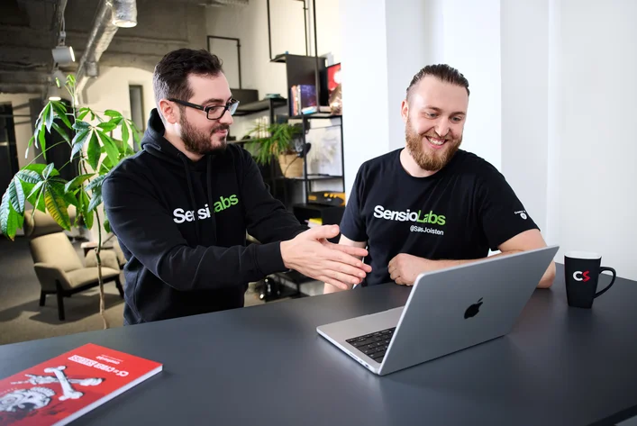 Dwóch programistów w koszulkach SensioLabs wspólnie omawia coś przy laptopie w biurze