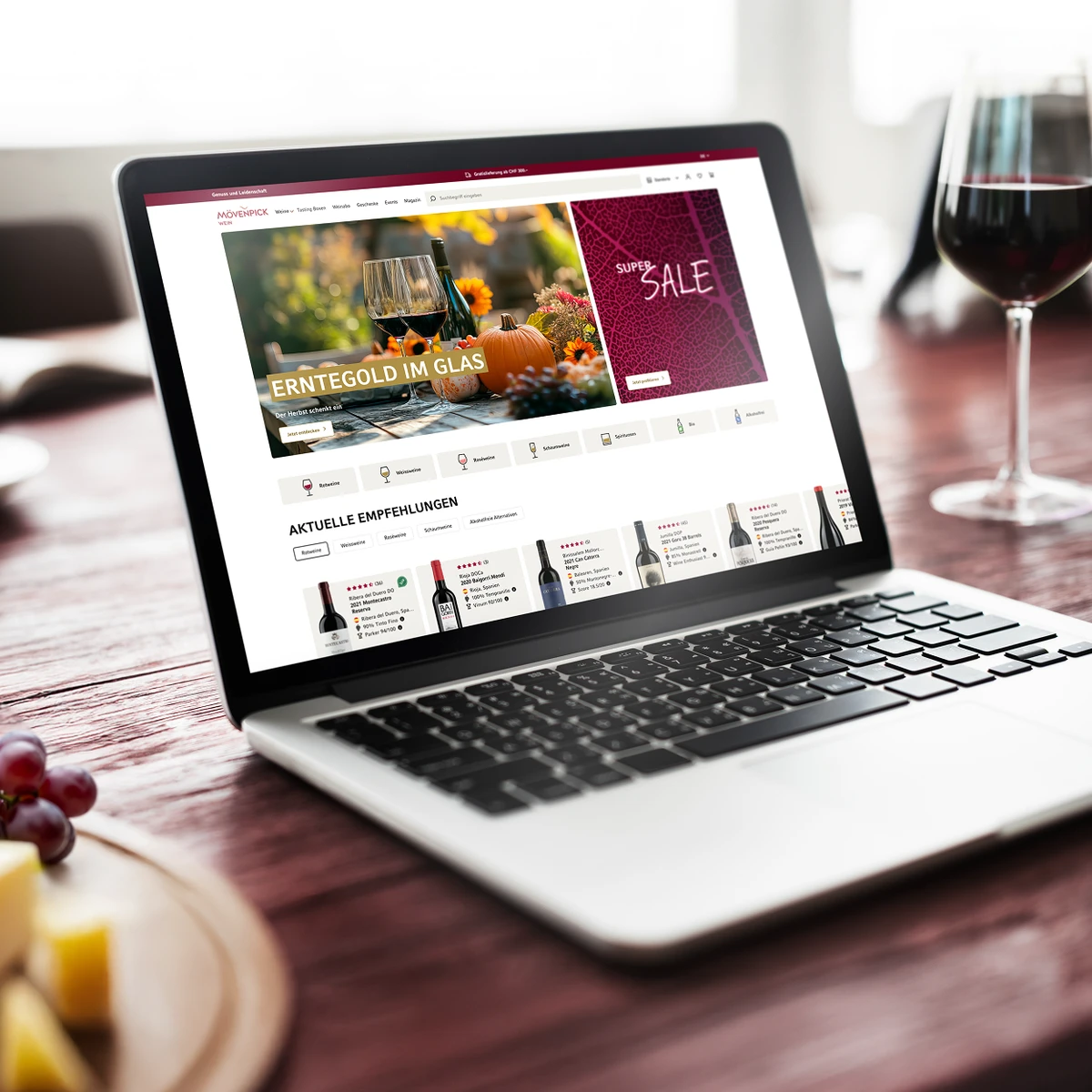 Podgląd strony e-commerce Mövenpick Wein na laptopie prezentujący jesienne promocje i aktualne rekomendacje win na tle klimatycznego wnętrza restauracyjnego z kieliszkiem czerwonego wina, winogronami i serem