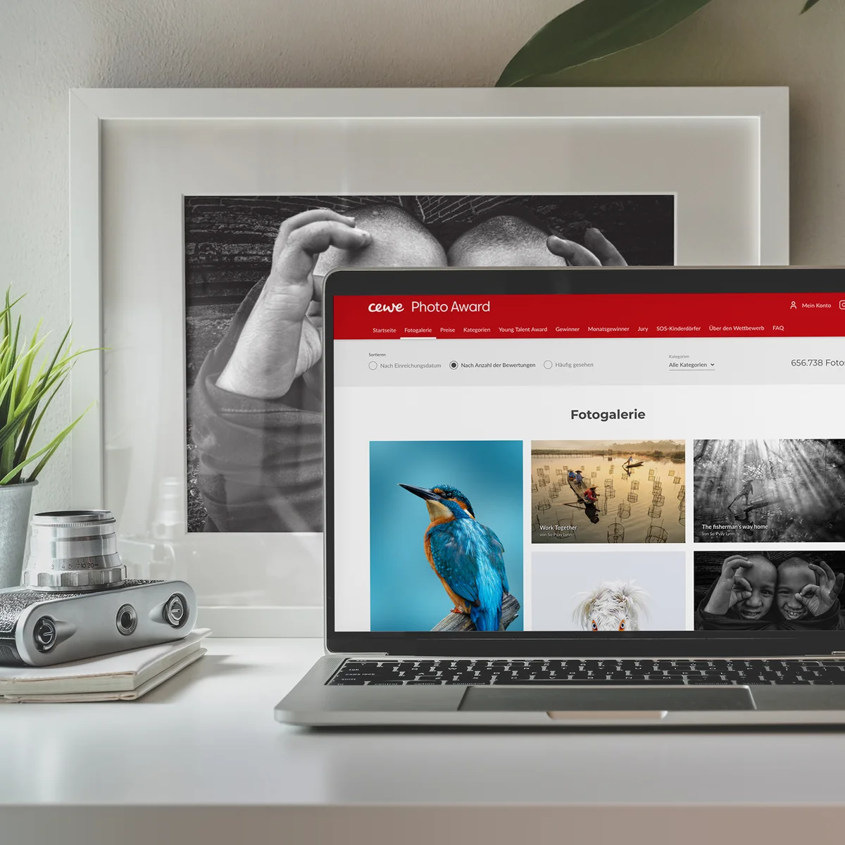 Podgląd strony e-commerce CEWE na laptopie prezentujący galerię „CEWE Photo Award” z różnymi nagrodzonymi fotografiami na tle biurka z oprawionym czarno-białym zdjęciem i klasycznym aparatem fotograficznym