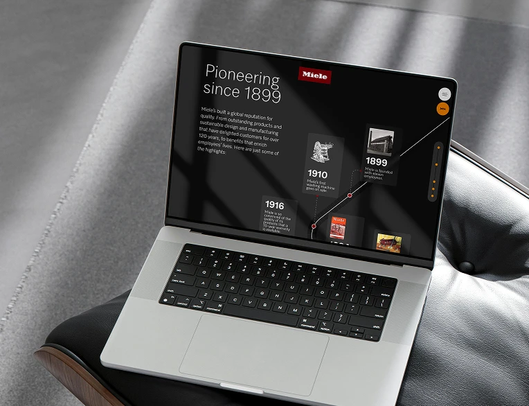 Podgląd strony e-commerce Miele X na laptopie prezentujący linię czasu historii marki „Pioneering since 1899” z kamieniami milowymi, takimi jak pierwsza pralka z 1910 roku, na ciemnym tle w luksusowym wnętrzu