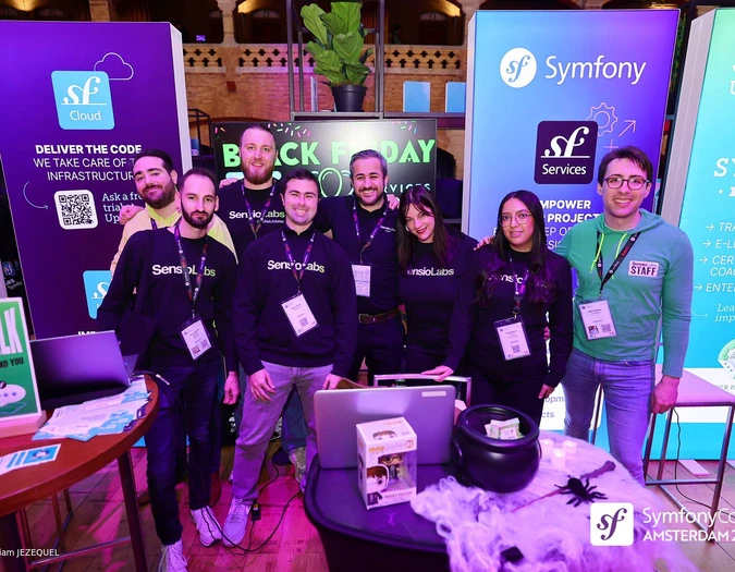 Grupa osób na stoisku SymfonyCon Amsterdam 2025 z różnymi banerami firmowymi