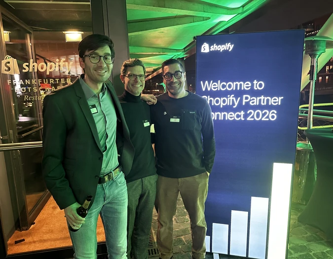 Trzech mężczyzn pozuje przy wejściu na wydarzenie Shopify obok niebieskiej tablicy „Welcome to Shopify Partner Connect 2026”